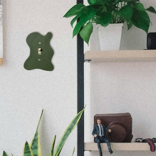 A dark green, gourd-shaped wall hook with a built-in single light switch is mounted on a textured off-white wall. The hook is styled in a home environment next to a wooden shelf holding a potted green plant and a miniature figure.