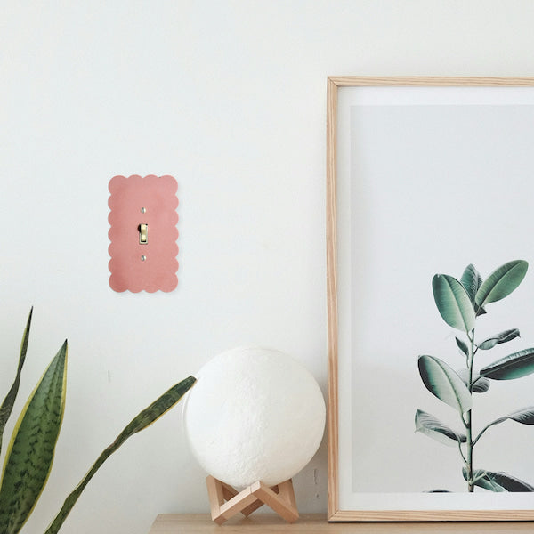A pink scalloped metal double light switch plate mounted on a white wall, styled with a white spherical lamp on a wooden stand and a framed botanical print.