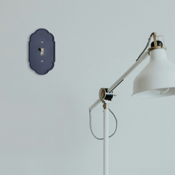 A navy blue decorative wall plate with a single toggle switch, mounted on a light gray wall next to a white floor lamp.