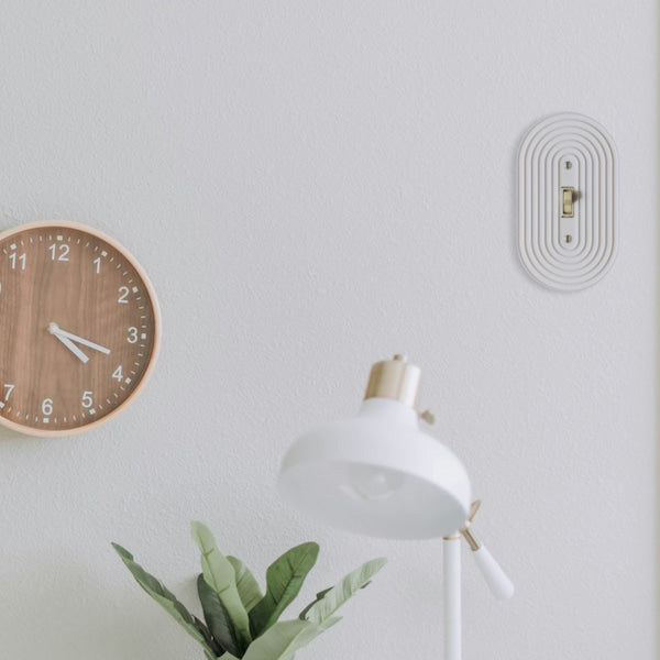 A white wall-mounted oval switch plate with concentric ridges and a small gold-toned center detail, shown on a light gray wall above a white desk lamp and next to a wooden wall clock.