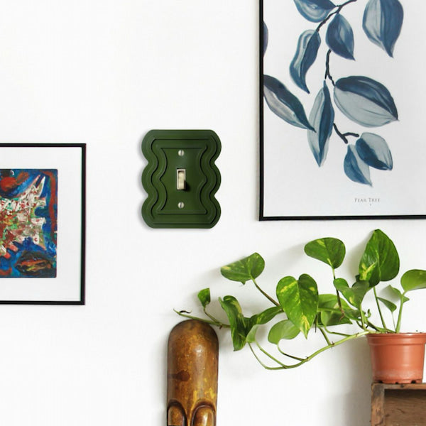 An olive green wavy double light switch plate mounted on a white wall, styled in a home interior next to framed art, a potted green plant, and a wooden sculpture.
